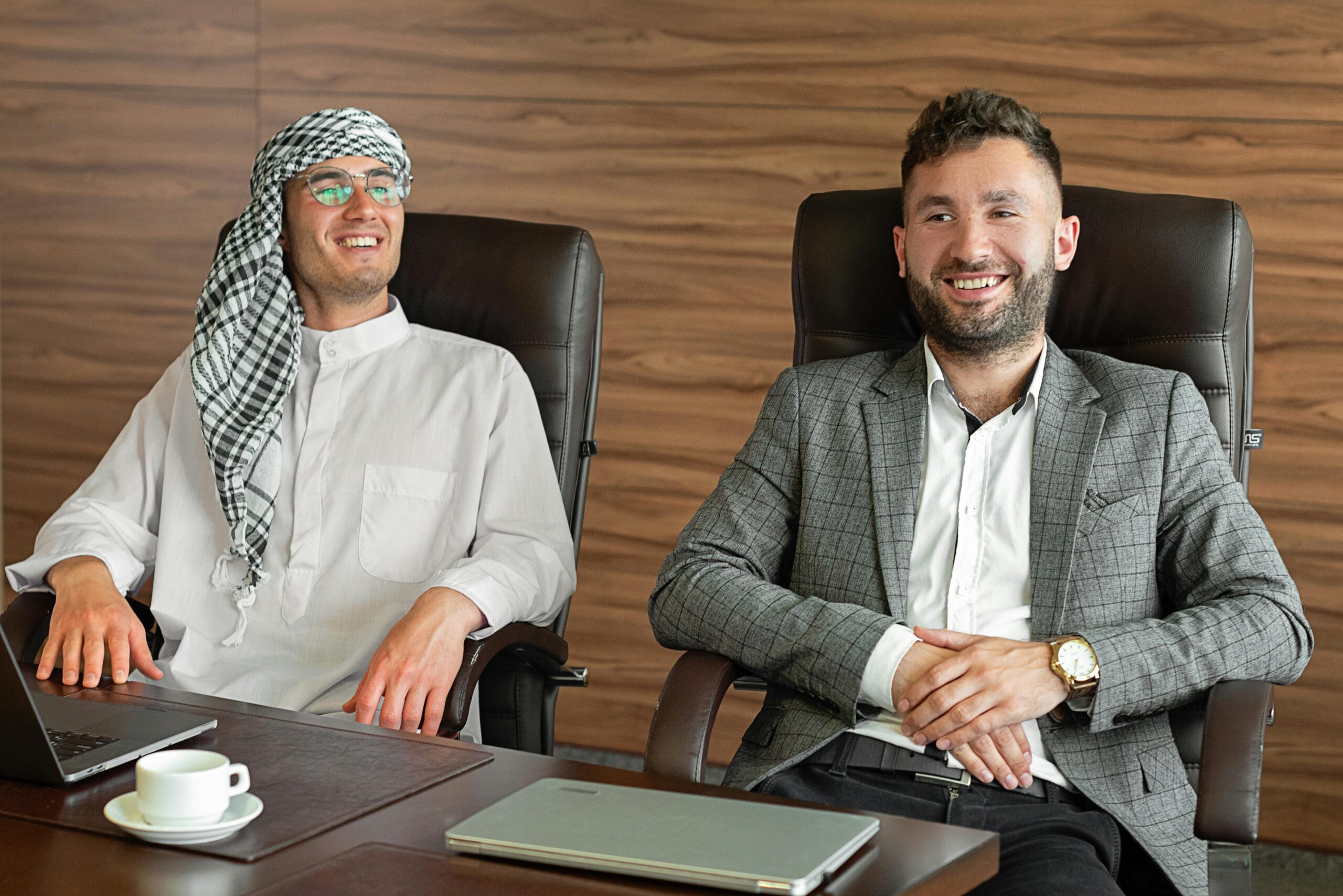 Two businessmen in traditional and modern attire sharing a laugh during a meeting in a stylish office.